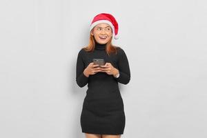 jovem asiática sorridente com chapéu de Papai Noel segurando um telefone celular e olhando para longe, isolada sobre fundo branco foto