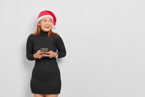 Mulher asiática jovem sorridente com chapéu de Papai Noel segurando um telefone celular e olhando para o espaço da cópia isolado sobre o fundo branco