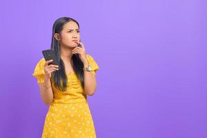 retrato de uma jovem mulher asiática pensativa usando um telefone celular e olhando para longe no fundo roxo foto