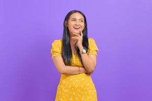 retrato de uma jovem asiática sorridente, tocando o queixo e desviando o olhar no fundo roxo foto