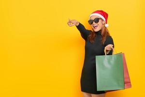 alegre jovem asiática com chapéu de Papai Noel e óculos de sol segurando sacolas de compras, apontando o dedo para o espaço da cópia isolado sobre fundo amarelo