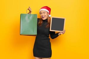 Mulher asiática jovem sorridente com chapéu de Papai Noel segurando sacolas de compras e um quadro-negro isolado sobre fundo amarelo