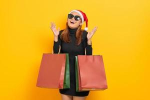 alegre jovem asiática com chapéu de Papai Noel e óculos escuros segurando sacolas de compras e comemorar a sorte isolada sobre fundo amarelo foto