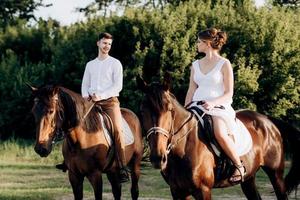 uma garota com um vestido de verão branco e um cara com uma camisa branca em uma caminhada com cavalos marrons foto