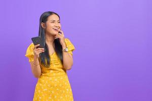 retrato de uma jovem asiática sorridente, usando um telefone celular e olhando para longe no fundo roxo foto