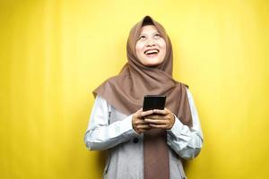 bela jovem muçulmana asiática chocada, surpresa, expressão uau, mão segurando o smartphone, olhando para cima, isolado em fundo amarelo foto