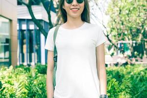 Hipster asiático menina cabelos castanhos compridos em t-shirt branca em branco está de pé no meio da rua. uma mulher com roupas de rua está de pé sobre um fundo urbano verde. mock up vazio espaço para texto ou design. foto