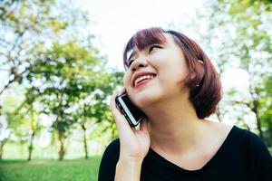 linda mulher asiática sorri e falando no celular enquanto está sentado no parque, dia de primavera. mulher asiática usando telefone inteligente com sensação de relaxamento e rosto sorridente. conceitos de estilo de vida e tecnologia. foto