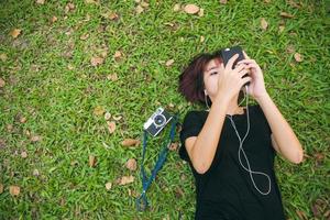 jovem mulher asiática deitado na grama verde, ouvindo música no parque, com uma emoção fria. jovem relaxando na grama com a câmera ao lado. atividade ao ar livre no conceito de parque.