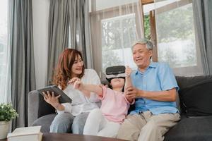 os avós e a neta asiáticos que usam a realidade virtual e o tablet jogam em casa. sênior chinês, vovô e vovó felizes relaxam com uma jovem deitada no sofá na sala de estar conceito. foto