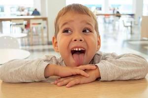 menino lindo e alegre faz caretas em um café, em uma mesa na praça de alimentação do shopping. Shopping. foto