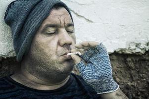 um mendigo com luvas e cara suja fuma um cigarro. pobreza, fome, desemprego.