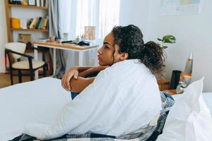vista lateral de uma mulher afro-americana pensativa, enrolada em um cobertor na cama