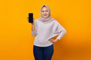 Mulher asiática jovem feliz mostrando a tela em branco do celular isolada sobre um fundo amarelo foto