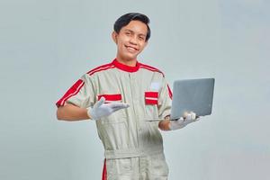 Cheeful bonito jovem mecânico vestindo uniforme apontando para o laptop com a palma da mão no fundo cinza foto