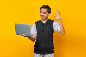 alegre bonito homem asiático segurando laptop e mostrando um gesto ok sobre fundo amarelo