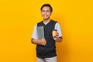 sorridente jovem bonito segurando laptop e segurando uma xícara de café sobre fundo amarelo foto