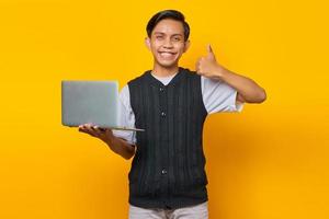 bonito homem asiático segurando laptop sorrindo e mostrando os polegares para cima sinal sobre fundo amarelo