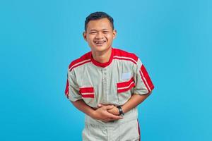retrato de um jovem mecânico asiático, mostrando gargalhadas com as mãos na barriga, sobre fundo azul