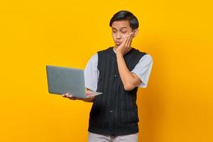 retrato de um jovem asiático sorridente segurando um laptop com a mão na bochecha em fundo amarelo