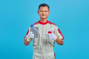 retrato de um jovem mecânico asiático sorridente, apontando para uma garrafa de plástico de óleo de motor com o dedo isolado sobre fundo azul