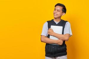 retrato de um homem alegre asiático carregando laptop e olhando para o espaço vazio sobre fundo amarelo