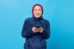 mulher asiática jovem e alegre usando o smartphone e olhando para a câmera sobre fundo azul