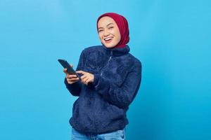 mulher asiática jovem e alegre usando o smartphone e olhando para a câmera sobre fundo azul