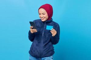 alegre jovem asiática segurando um telefone celular e mostrando um cartão de crédito sobre fundo azul