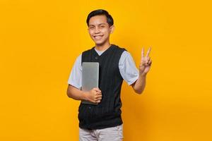 bonito homem asiático segurando laptop, sorrindo e mostrando o símbolo da paz sobre fundo amarelo foto