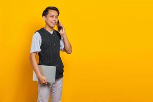 jovem empresário bonito falando em um smartphone e segurando um laptop sobre fundo amarelo