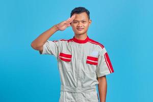 retrato de um jovem mecânico asiático sorridente, fazendo sinal de saudação com a palma da mão isolada sobre fundo azul.