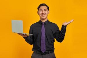 retrato de um jovem empresário asiático sorridente, mostrando as palmas das mãos e segurando o laptop sobre fundo amarelo foto