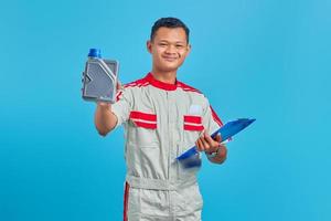 retrato de um jovem mecânico asiático sorrindo, segurando a prancheta e mostrando uma garrafa de plástico de óleo de motor na mão sobre fundo azul foto
