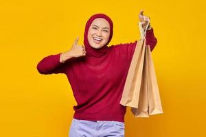 linda mulher asiática mostrando algumas sacolas de compras e dando os polegares para cima com uma cara sorridente no fundo amarelo