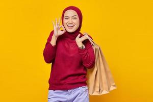retrato de uma mulher asiática alegre segurando sacolas de compras e fazendo sinal de ok com o dedo sobre fundo amarelo