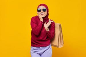 mulher asiática jovem feliz com a mão nas bochechas e segurando sacolas de compras no fundo amarelo