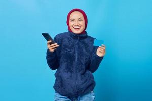 Retrato de uma jovem asiática sorridente, segurando um telefone celular e mostrando o cartão de crédito sobre fundo azul