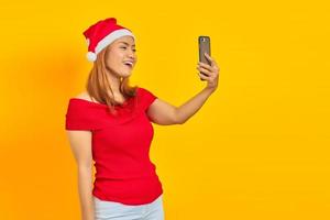 jovem asiática alegre usando chapéu de natal fazendo videochamada em fundo amarelo