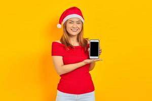 jovem asiática sorridente com chapéu de Papai Noel mostrando tela em branco do celular isolado sobre fundo amarelo foto