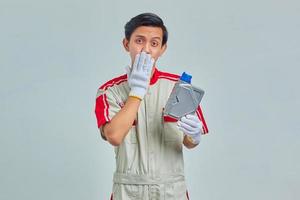 retrato de homem bonito surpreso usando uniforme de mecânico, segurando uma garrafa de plástico de óleo de motor e cobrindo a boca com as mãos no fundo cinza foto