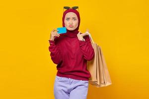 jovem asiática chocada segurando um cartão de crédito e sacolas de compras em fundo amarelo