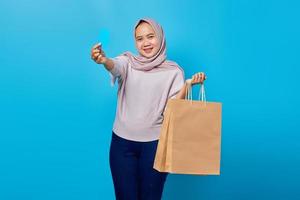 retrato de uma mulher asiática alegre segurando uma sacola de compras e mostrando o cartão de crédito sobre fundo azul