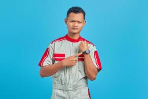 retrato de um jovem mecânico asiático zangado, mostrando as horas no relógio de pulso sobre o fundo azul foto