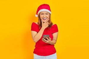 alegre jovem asiática usando chapéu de Natal usando smartphone com o dedo no queixo sobre fundo amarelo foto