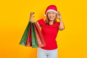 jovem mulher asiática sorridente segurando sacolas de compras e coçando a cabeça com a mão em fundo amarelo foto