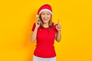 alegre jovem asiática usando chapéu de natal falando no celular e apontando o dedo para cima com uma ideia sobre fundo amarelo foto