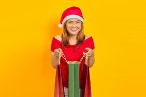 jovem mulher asiática sorridente com chapéu de Papai Noel abrindo a sacola de compras e olhando para a câmera sobre fundo amarelo
