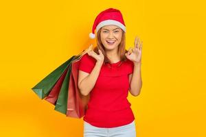 sorridente jovem mulher asiática com chapéu de Papai Noel segurando sacolas de compras e gesticulando um sinal de ok isolado sobre fundo amarelo