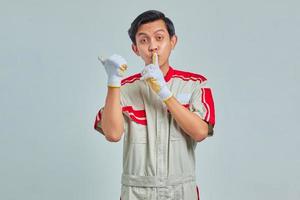 homem bonito usando uniforme de mecânico sinalizando para silenciar e apontando para o lado em fundo cinza foto